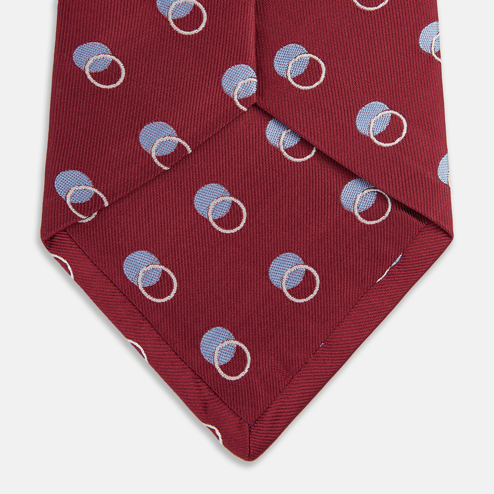 The Turnbull & Asser Red Multi Circle Silk Tie features small blue and white rings on red silk, making it ideal for pairing with bold shirts or adding vibrant flair to summer looks. Shown rolled at one end against a plain white background.