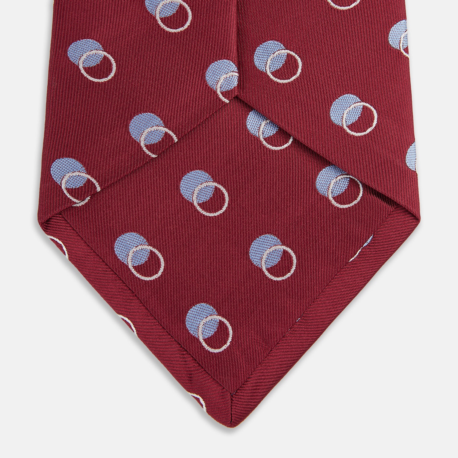 View 4 of product The Turnbull & Asser Red Multi Circle Silk Tie features small blue and white rings on red silk, making it ideal for pairing with bold shirts or adding vibrant flair to summer looks. Shown rolled at one end against a plain white background.