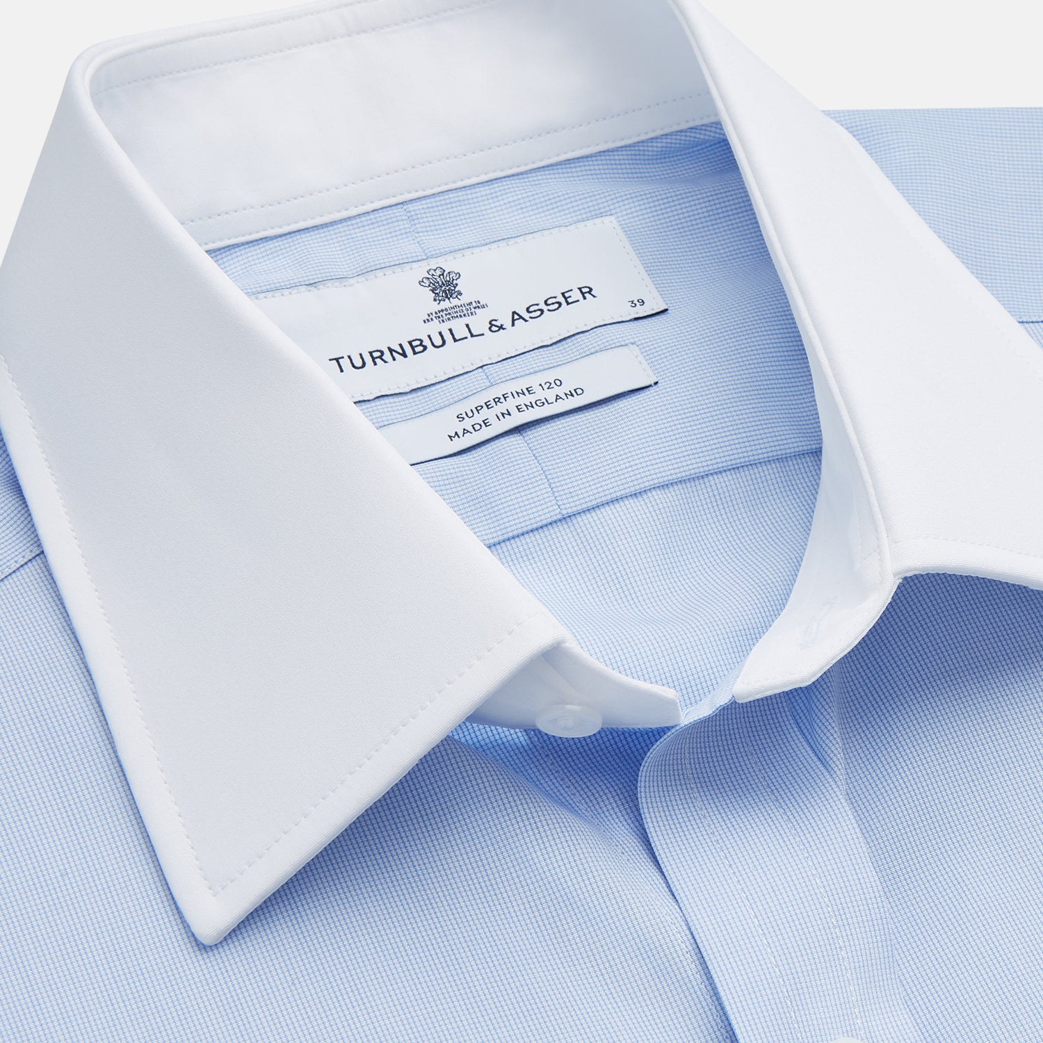 View 3 of product A man stands against a tan background wearing the Turnbull & Asser Mid Blue Micro-Check Regular Fit Shirt with White T&A Collar and Double Cuffs, paired with a brown patterned tie, dark trousers, and black dress shoes.