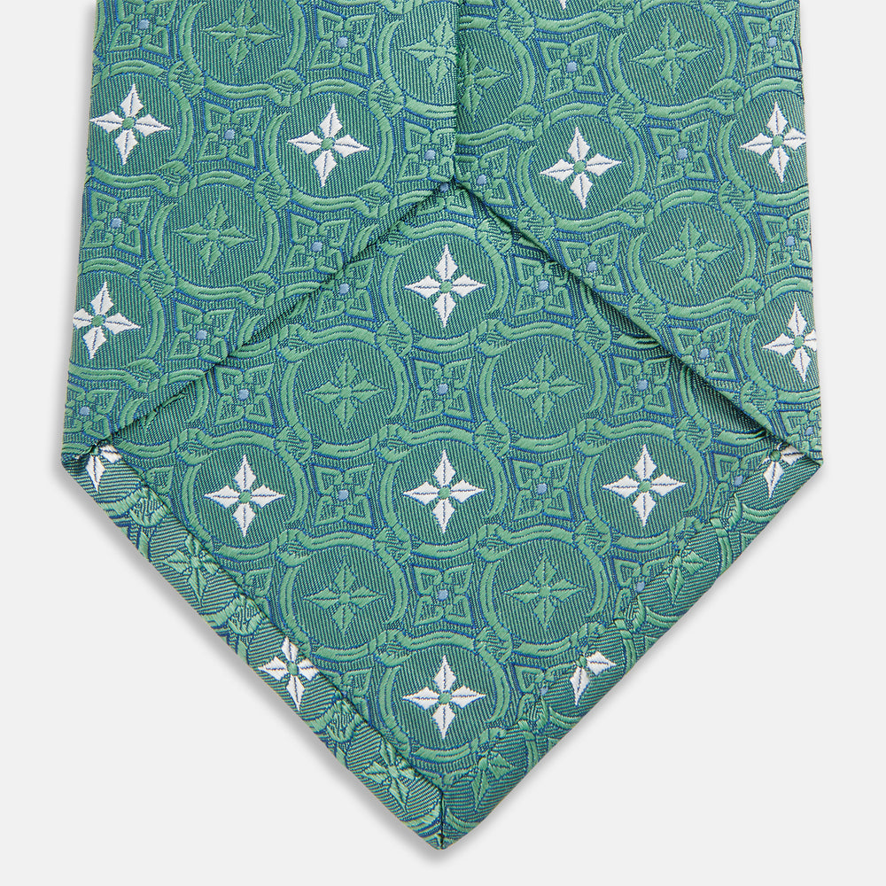 The Turnbull & Asser Green Multi Neat Silk Tie, handmade and featuring a white and light blue geometric pattern, is shown rolled up on a plain white background.
