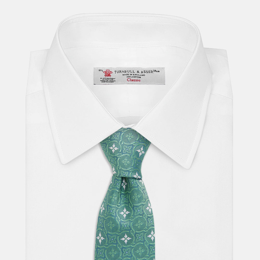 The Turnbull & Asser Green Multi Neat Silk Tie, handmade and featuring a white and light blue geometric pattern, is shown rolled up on a plain white background.