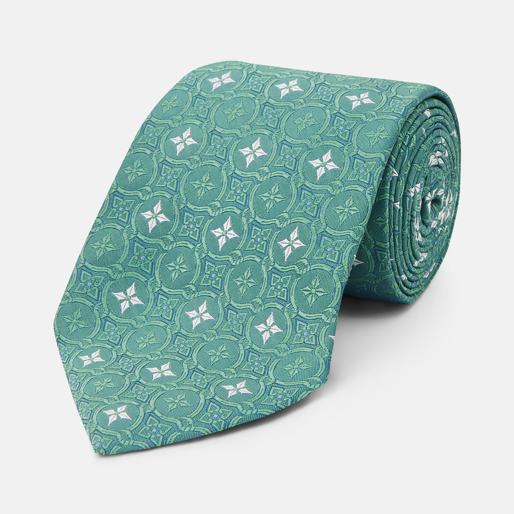 The Turnbull & Asser Green Multi Neat Silk Tie, handmade and featuring a white and light blue geometric pattern, is shown rolled up on a plain white background.