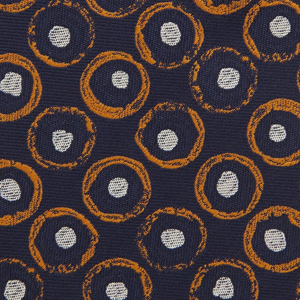 The Turnbull & Asser Orange Multi Circle Printed Silk Tie features navy blue silk with orange circles and white ovals, perfect for elevating any formal outfit. Shown on a plain white background.