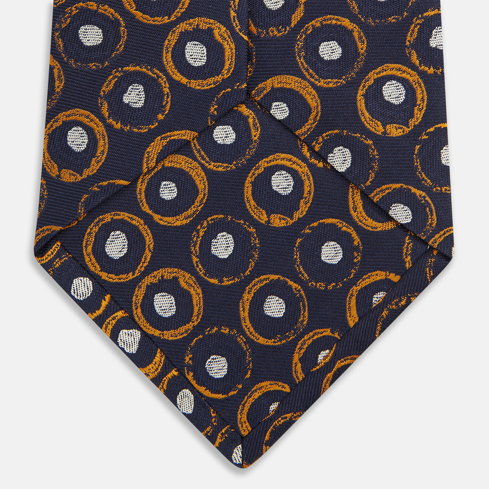 The Turnbull & Asser Orange Multi Circle Printed Silk Tie features navy blue silk with orange circles and white ovals, perfect for elevating any formal outfit. Shown on a plain white background.