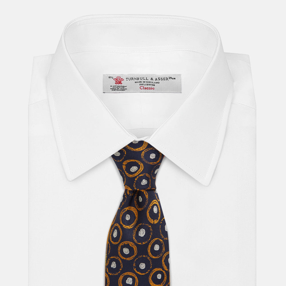 The Turnbull & Asser Orange Multi Circle Printed Silk Tie features navy blue silk with orange circles and white ovals, perfect for elevating any formal outfit. Shown on a plain white background.