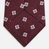 View 4 of product The Turnbull & Asser Burgundy Multi Geo Silk Tie features burgundy silk with blue, white, and yellow geometric designs—ideal for bold statement shirts. Shown rolled against a plain white background.