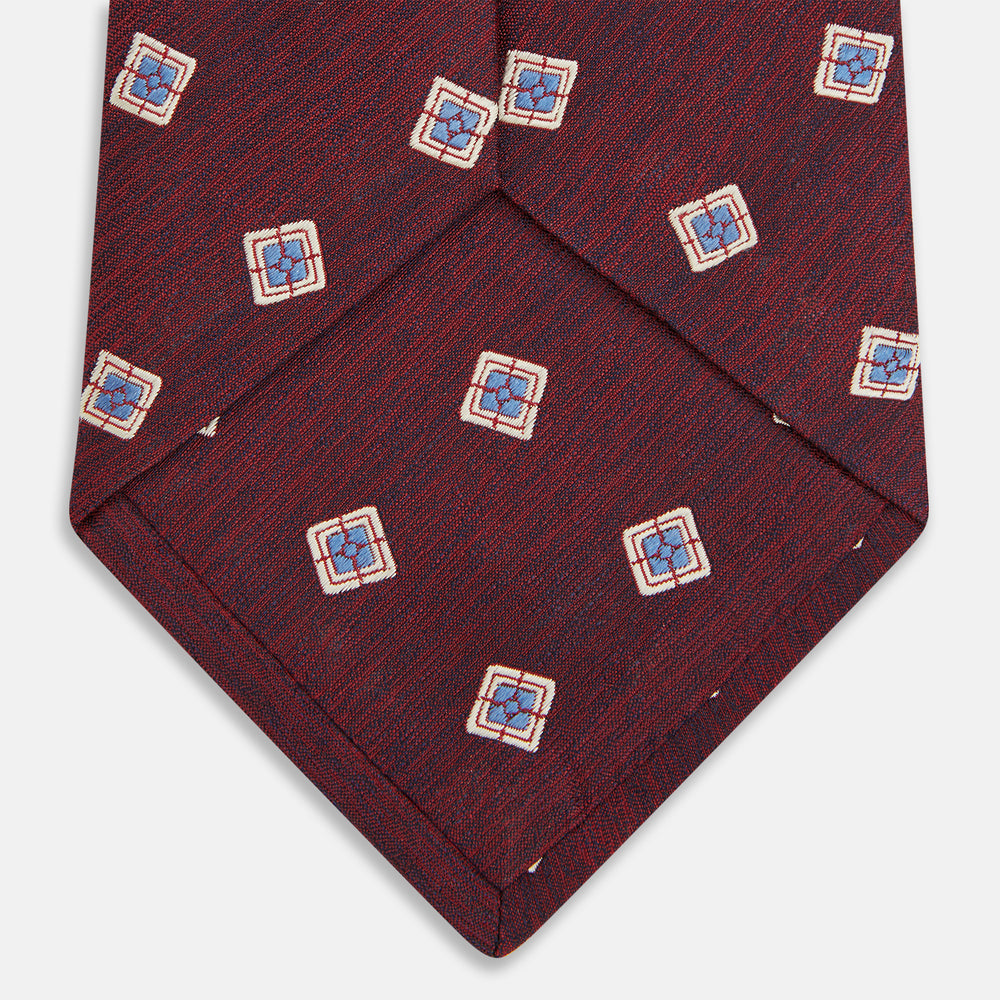 The Turnbull & Asser Burgundy Multi Geo Silk Tie features burgundy silk with blue, white, and yellow geometric designs—ideal for bold statement shirts. Shown rolled against a plain white background.