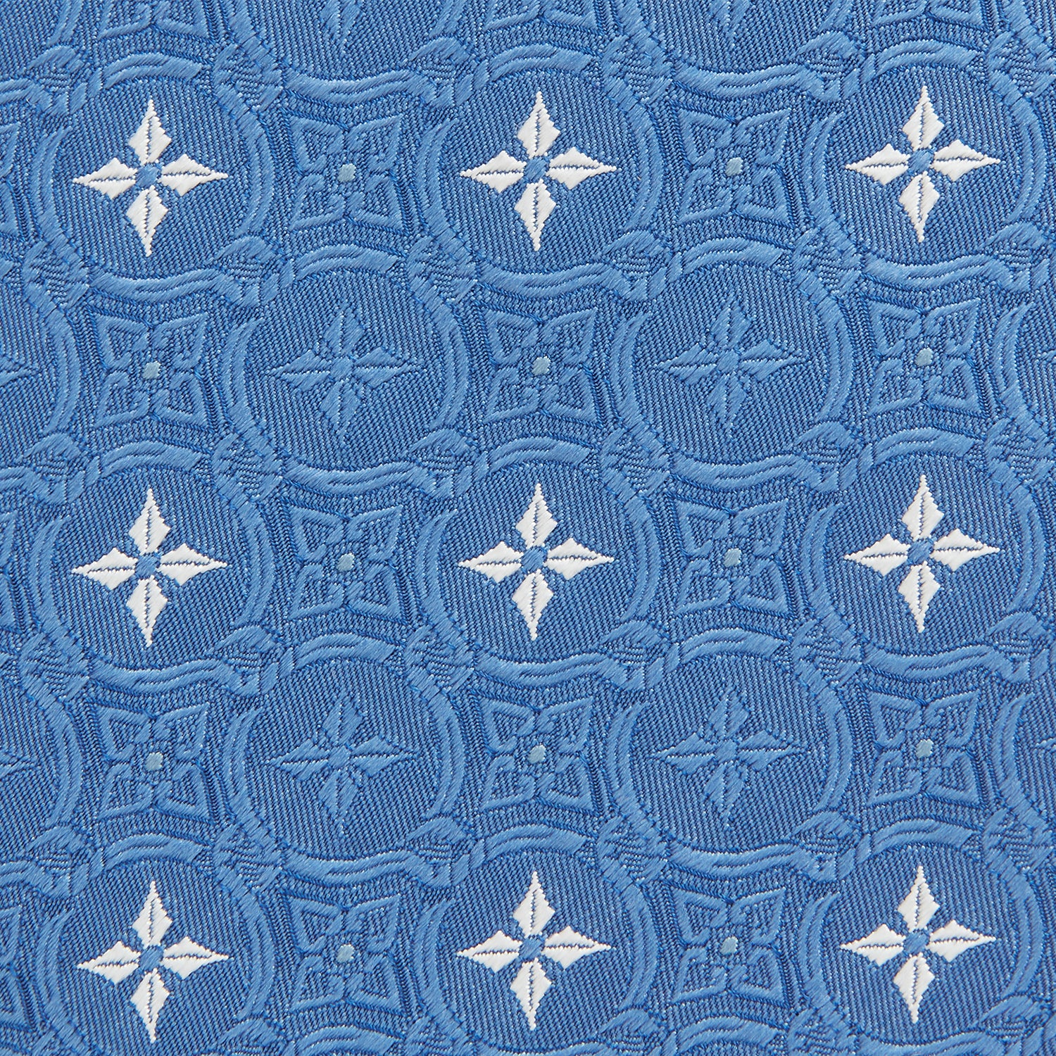 View 3 of product The Blue Multi Neat Silk Tie by Turnbull & Asser features a geometric pattern of white four-pointed shapes and circles, crafted in blue silk from the autumn-winter collection. Displayed rolled on a plain light background.