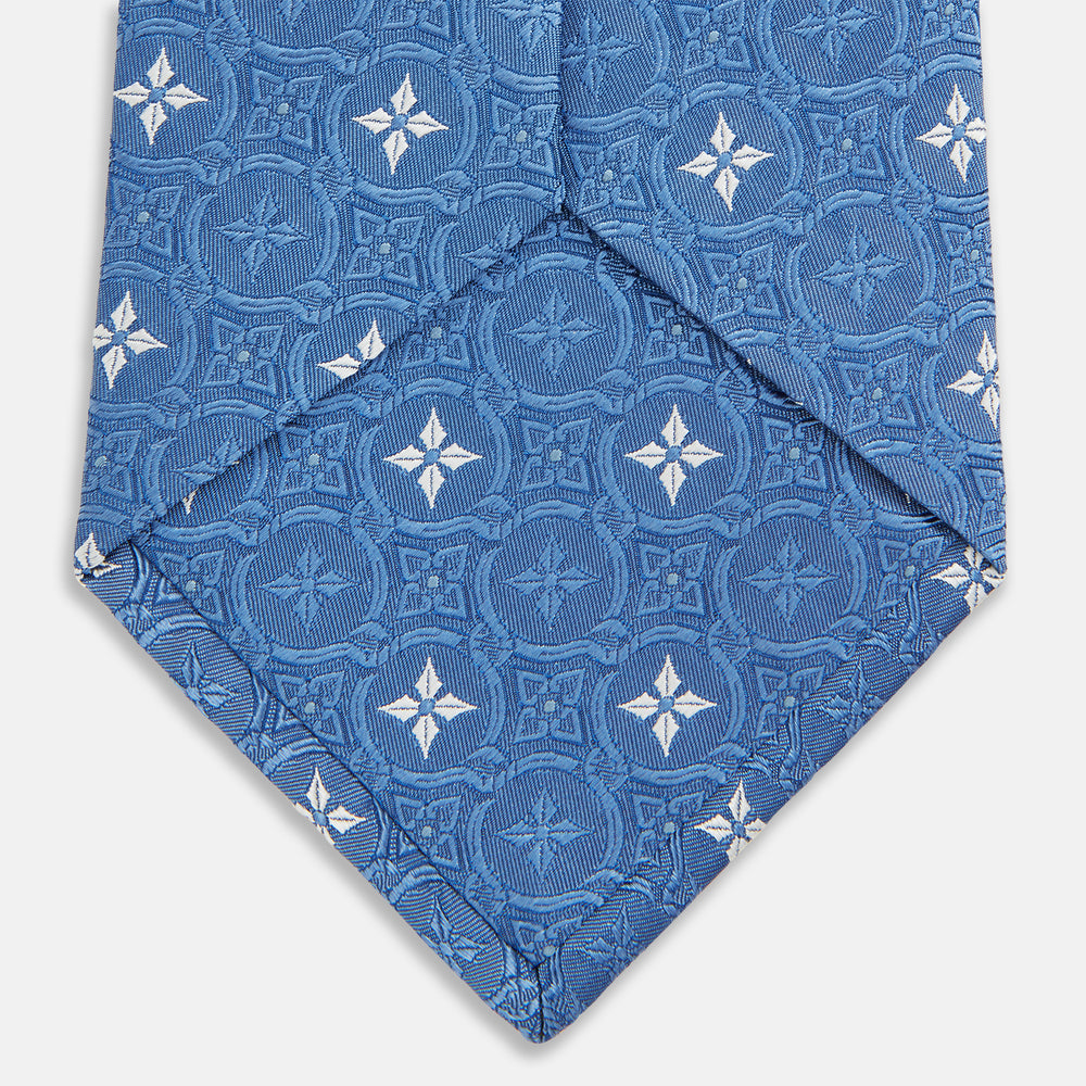 The Blue Multi Neat Silk Tie by Turnbull & Asser features a geometric pattern of white four-pointed shapes and circles, crafted in blue silk from the autumn-winter collection. Displayed rolled on a plain light background.