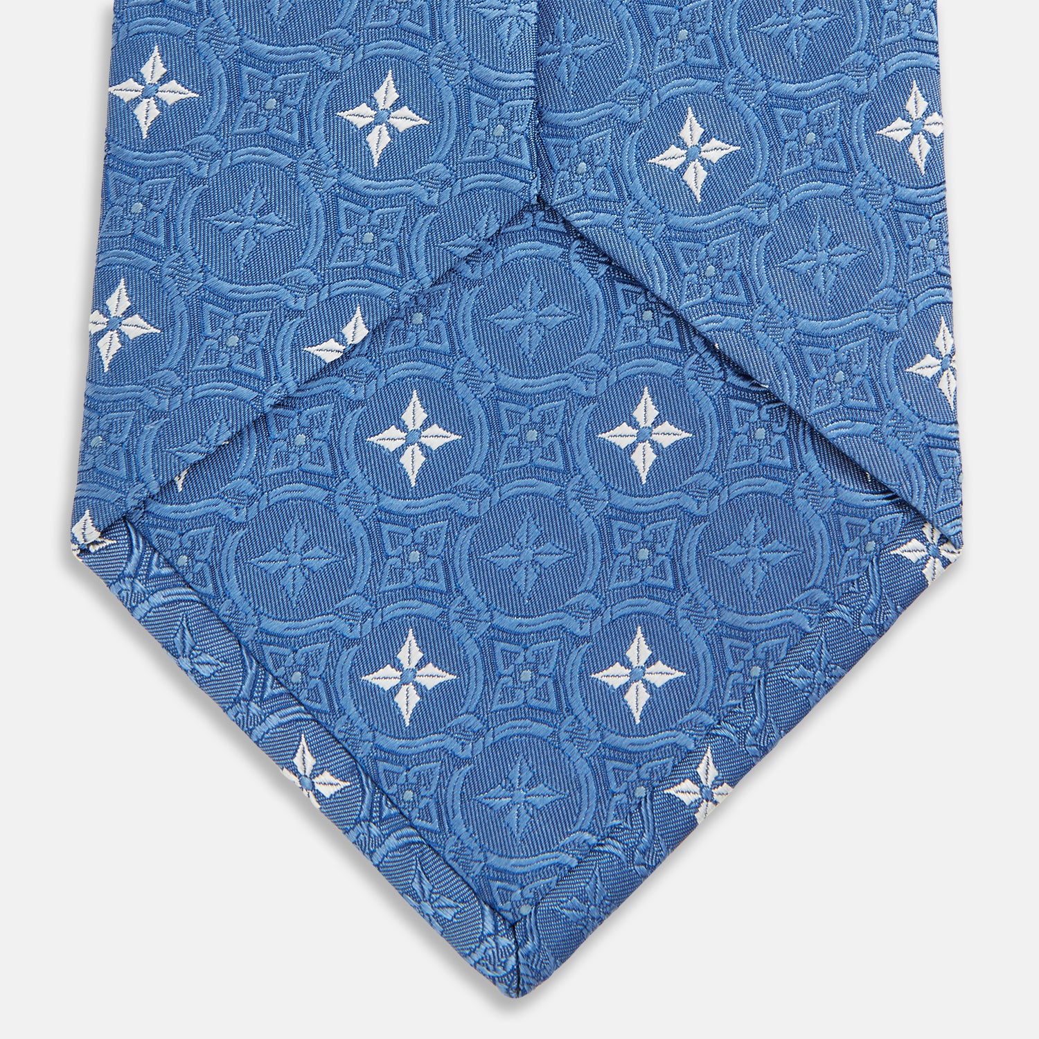 View 4 of product The Blue Multi Neat Silk Tie by Turnbull & Asser features a geometric pattern of white four-pointed shapes and circles, crafted in blue silk from the autumn-winter collection. Displayed rolled on a plain light background.