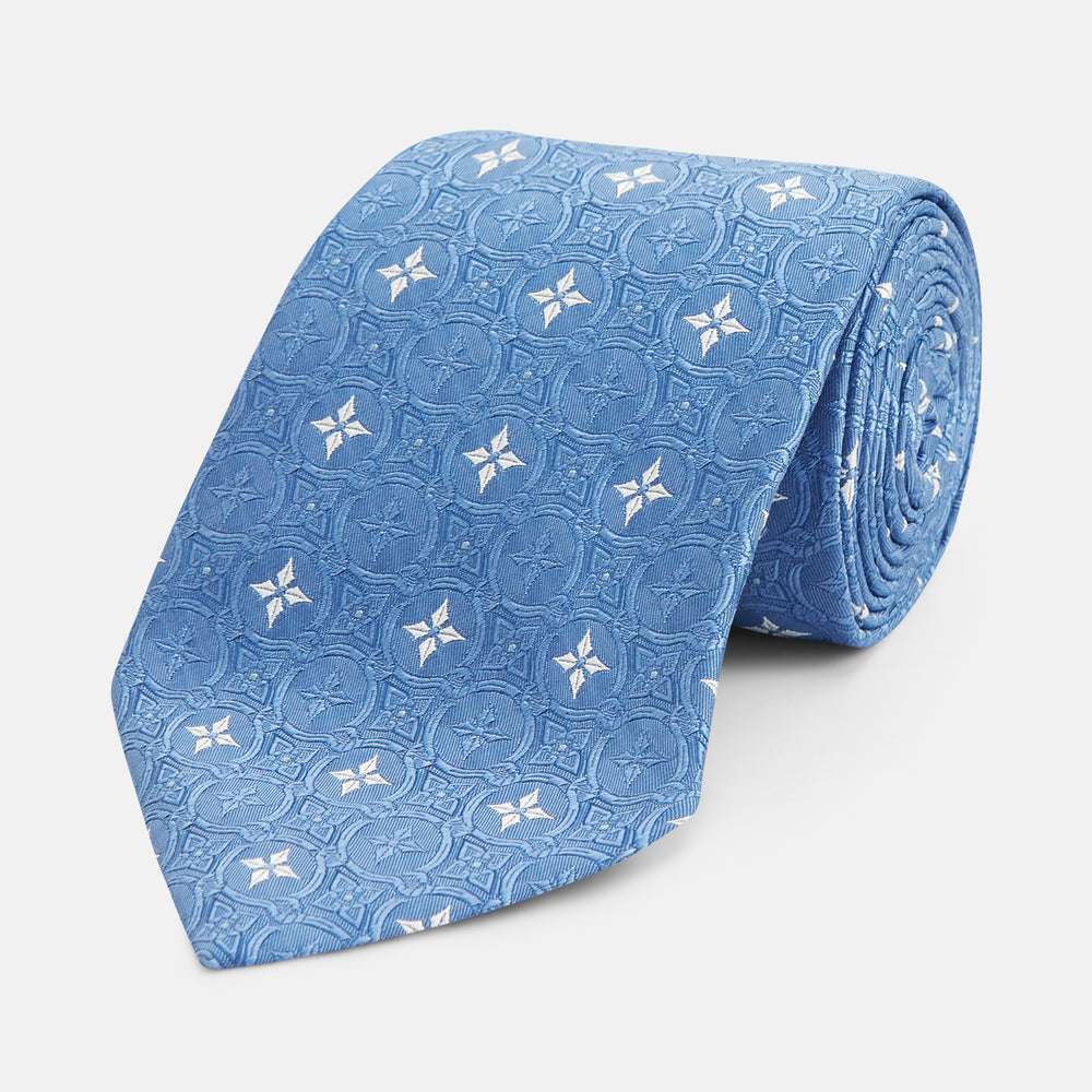 The Blue Multi Neat Silk Tie by Turnbull & Asser features a geometric pattern of white four-pointed shapes and circles, crafted in blue silk from the autumn-winter collection. Displayed rolled on a plain light background.
