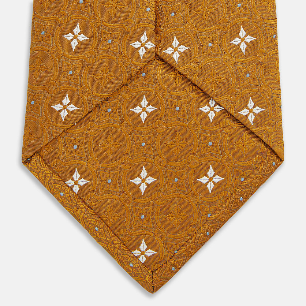 The Orange Multi Neat Silk Tie by Turnbull & Asser, from the autumn-winter collection, features a golden yellow geometric pattern on textured silk. Shown rolled on a white background, it pairs perfectly with bold signature shirts.
