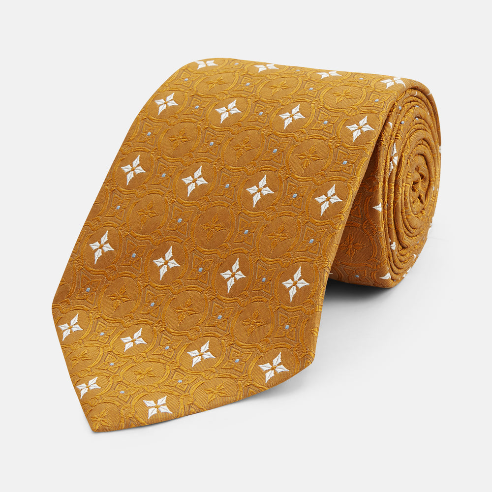 The Orange Multi Neat Silk Tie by Turnbull & Asser, from the autumn-winter collection, features a golden yellow geometric pattern on textured silk. Shown rolled on a white background, it pairs perfectly with bold signature shirts.
