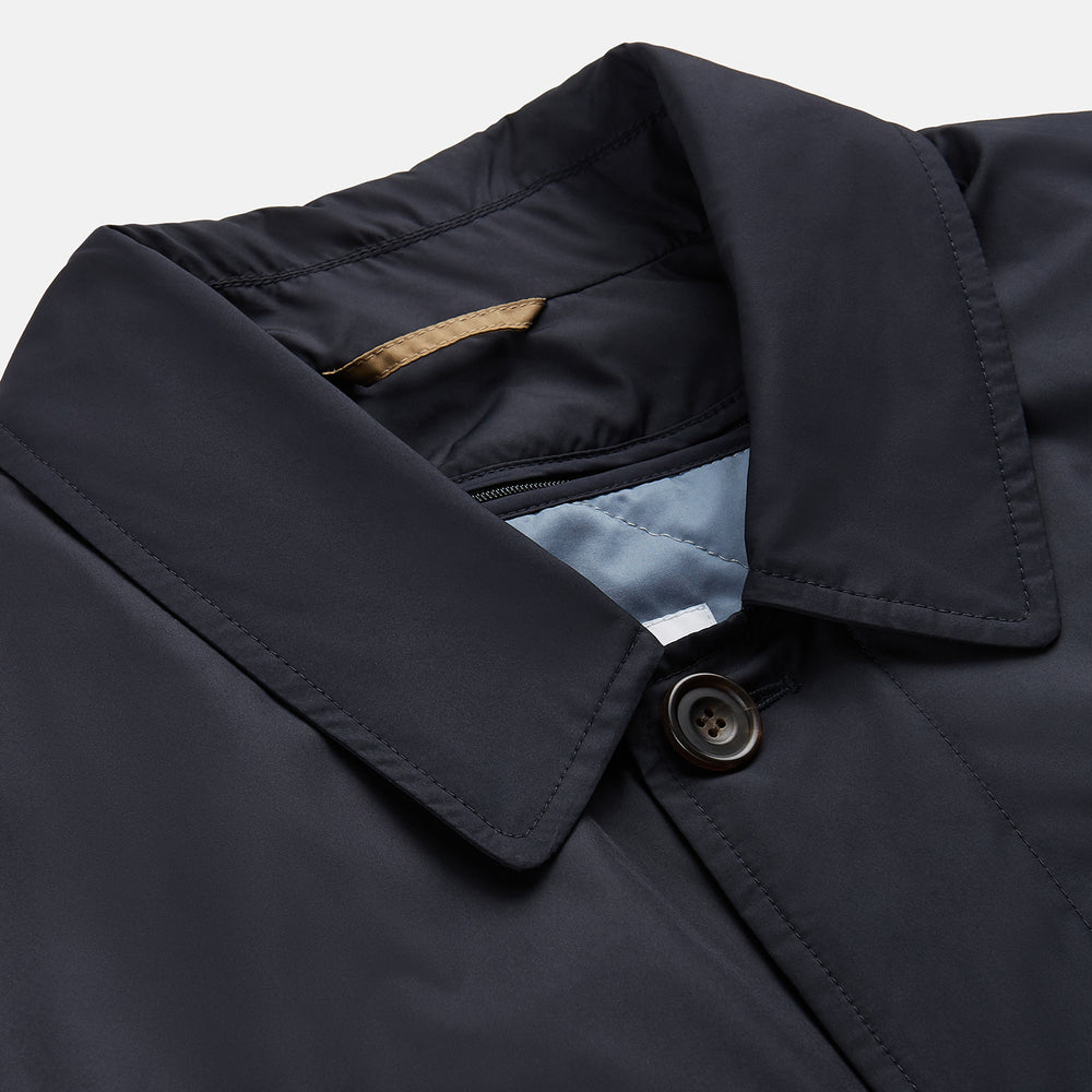 A man with brown hair stands outdoors, hands in his pockets, wearing a Turnbull & Asser Navy Sealup Quilted Liner Quintern Car Coat over a mustard cardigan. A glass barrier and blurred background are behind him.