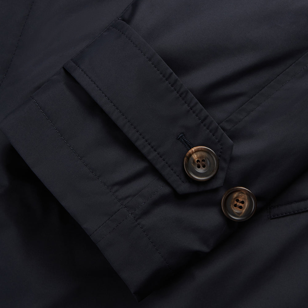 A man with brown hair stands outdoors, hands in his pockets, wearing a Turnbull & Asser Navy Sealup Quilted Liner Quintern Car Coat over a mustard cardigan. A glass barrier and blurred background are behind him.