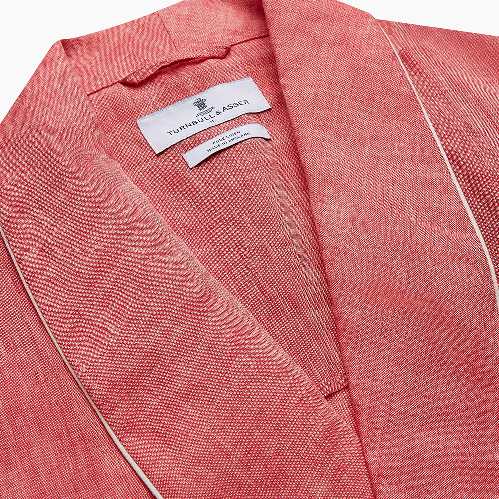 The Pink Linen Arnold Gown by Turnbull & Asser features white piping, long sleeves, a shawl collar, chest pocket, and a wrap-around belt tied at the waist, shown on a white background.