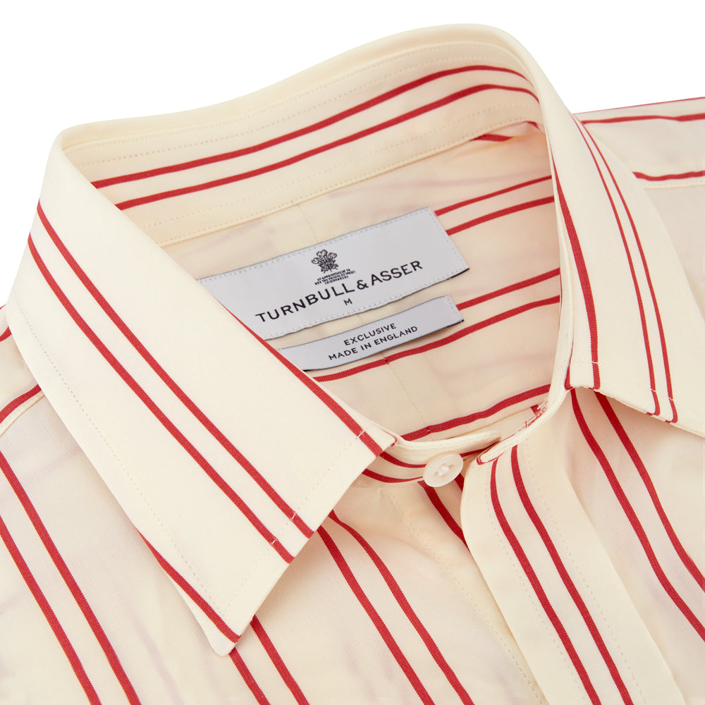 A man poses against a beige backdrop in the Turnbull & Asser Red and Off-White Stripe Cotton Weekend Fit Finch Shirt, styled with dark blue jeans and a brown belt for a timeless look. One hand rests in his pocket as he faces slightly to the side.