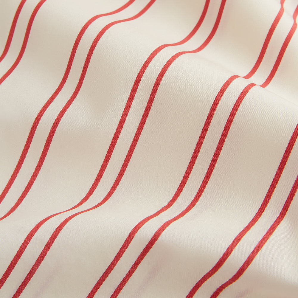 A man poses against a beige backdrop in the Turnbull & Asser Red and Off-White Stripe Cotton Weekend Fit Finch Shirt, styled with dark blue jeans and a brown belt for a timeless look. One hand rests in his pocket as he faces slightly to the side.