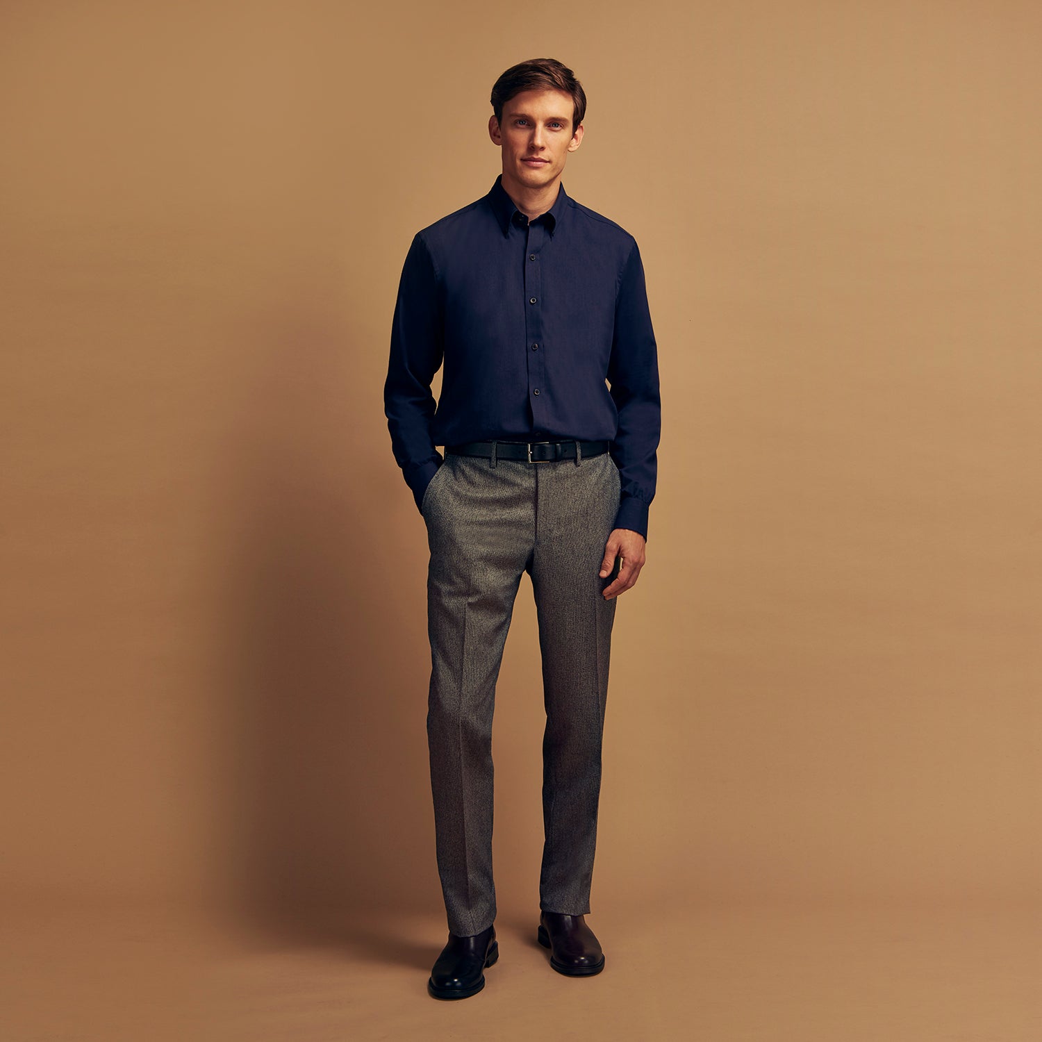 View 1 of product A man stands against a tan background, wearing the Turnbull & Asser Navy Wool Blend Weekend Fit Hayne Shirt tucked into gray dress pants with a black belt and shoes. He has short hair, a neutral expression, and his hands are in his pockets for a casual look.