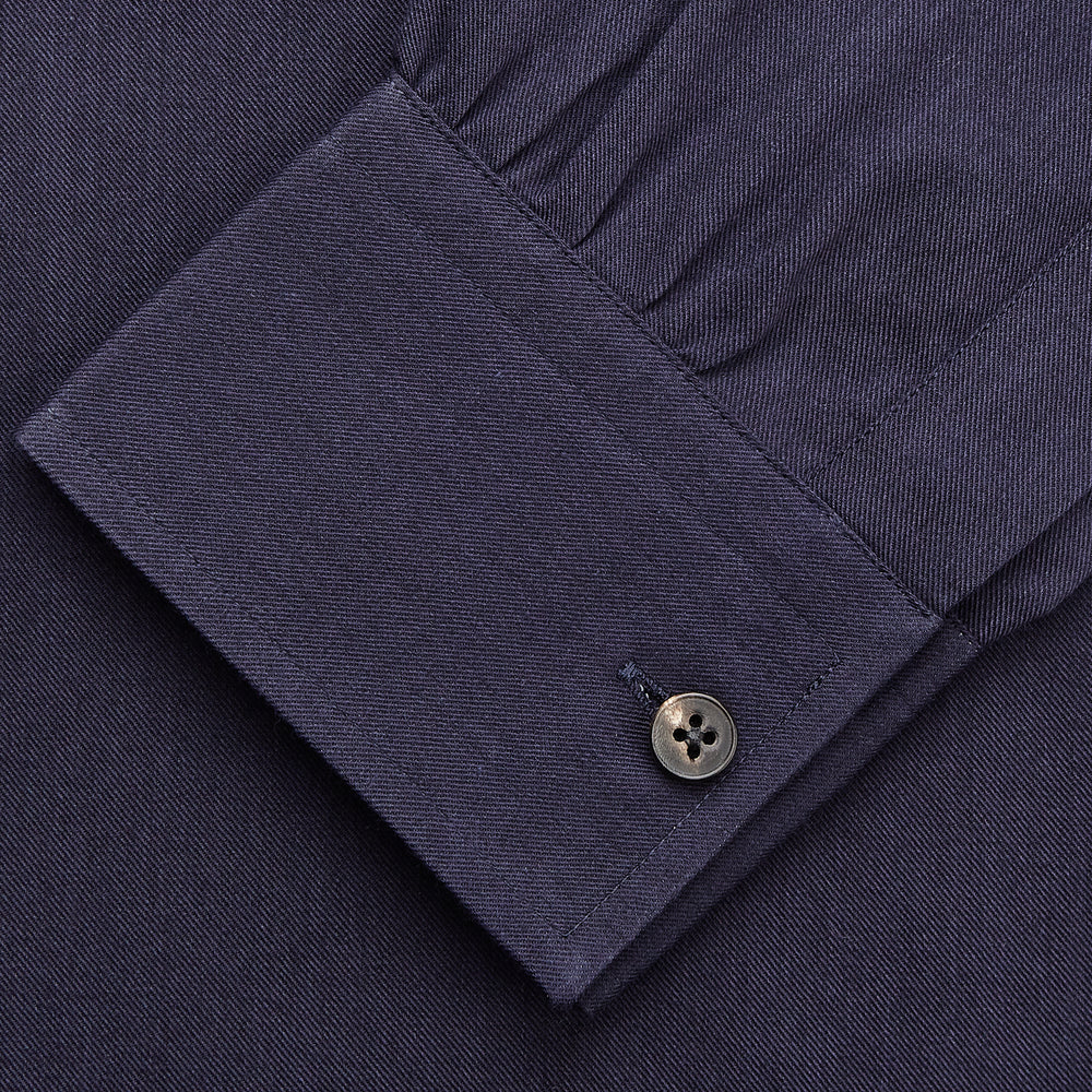 A man stands against a tan background, wearing the Turnbull & Asser Navy Wool Blend Weekend Fit Hayne Shirt tucked into gray dress pants with a black belt and shoes. He has short hair, a neutral expression, and his hands are in his pockets for a casual look.