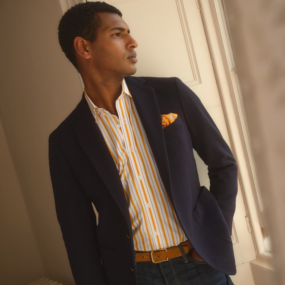 A man stands against a tan background wearing a Turnbull & Asser Orange Multi Stripe Regular Fit Shirt with T&A Collar and 3 Button Cuffs, paired with tan dress pants, brown belt, and brown shoes. He faces forward with a neutral expression.