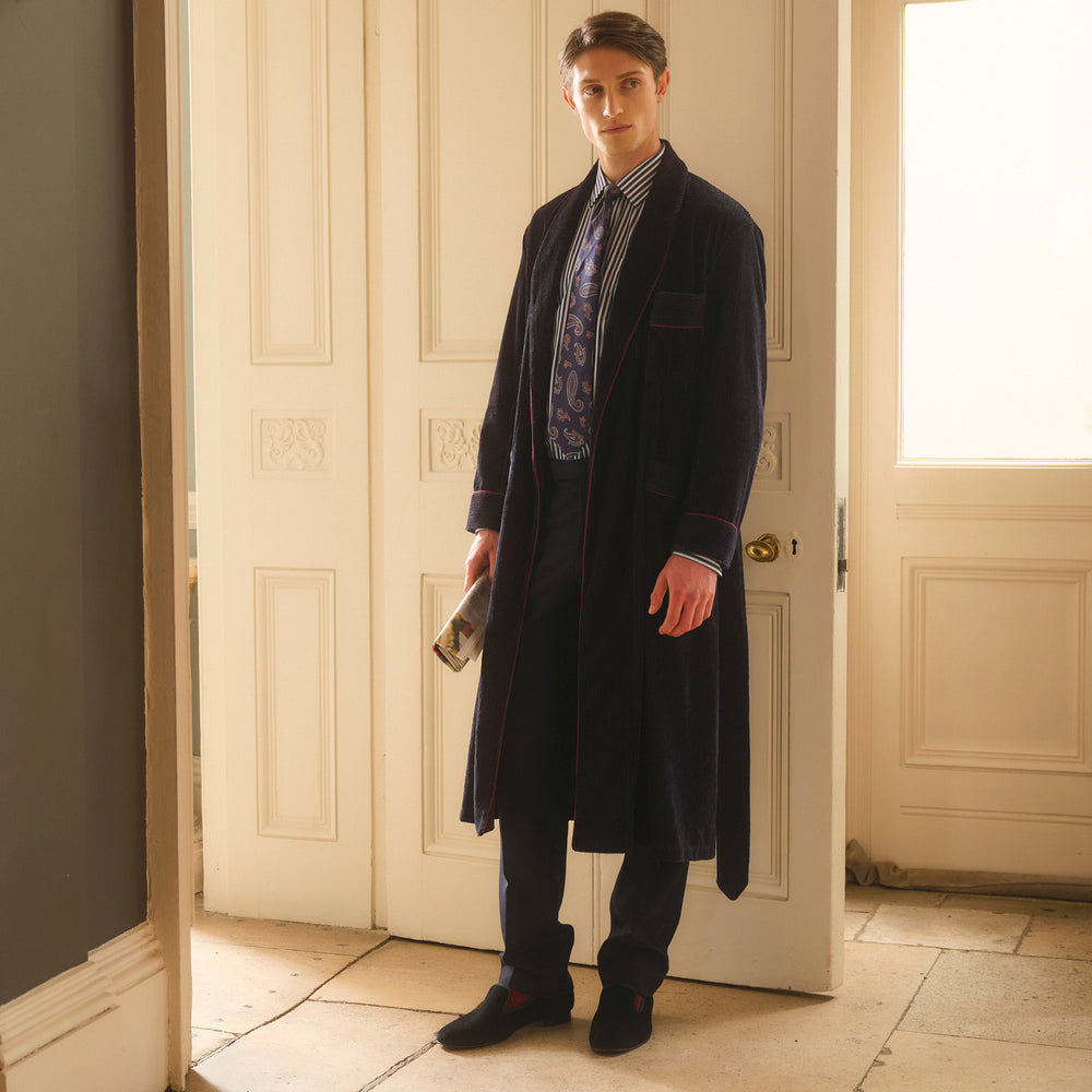 A man stands indoors before double doors, wearing the Turnbull & Asser Navy Linen & Cotton Cord Gown over a striped shirt and patterned tie, holding a book as soft natural light fills the scene.