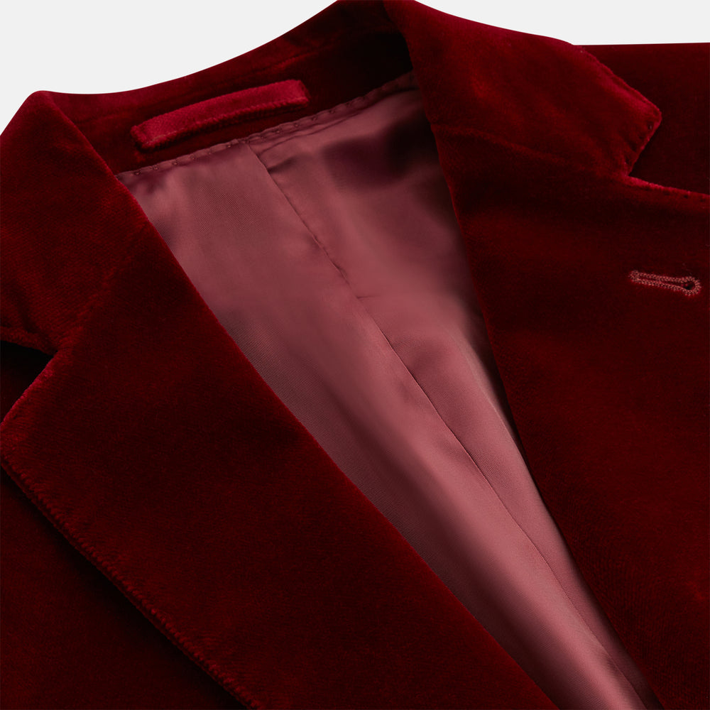 The Turnbull & Asser Wine Velvet Barrington Blazer, tailored in Italy, features a deep red hue, notched lapel, two front buttons, three pockets, and cuff button details. Shown against a plain white background.