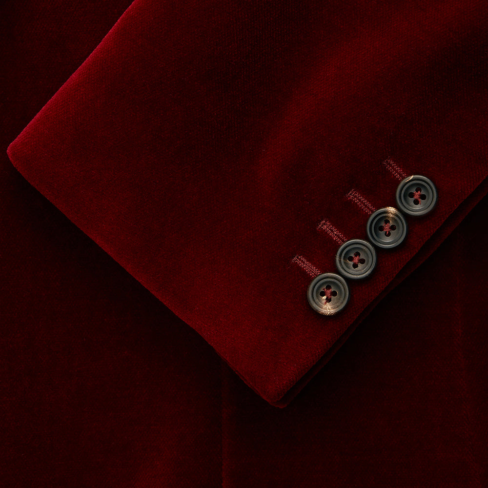 The Turnbull & Asser Wine Velvet Barrington Blazer, tailored in Italy, features a deep red hue, notched lapel, two front buttons, three pockets, and cuff button details. Shown against a plain white background.