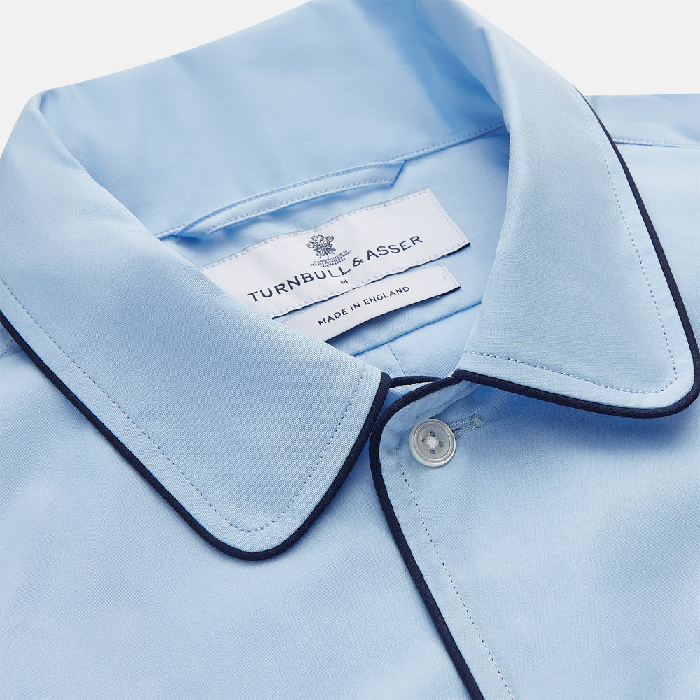 The Turnbull & Asser Light Blue Piped Cotton Nightshirt is a luxurious long-sleeve shirt dress with a pointed collar, black piping on the collar, cuffs, and chest pocket, button front closure, and curved hem.