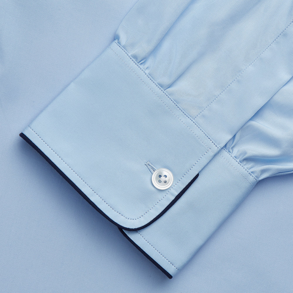 The Turnbull & Asser Light Blue Piped Cotton Nightshirt is a luxurious long-sleeve shirt dress with a pointed collar, black piping on the collar, cuffs, and chest pocket, button front closure, and curved hem.