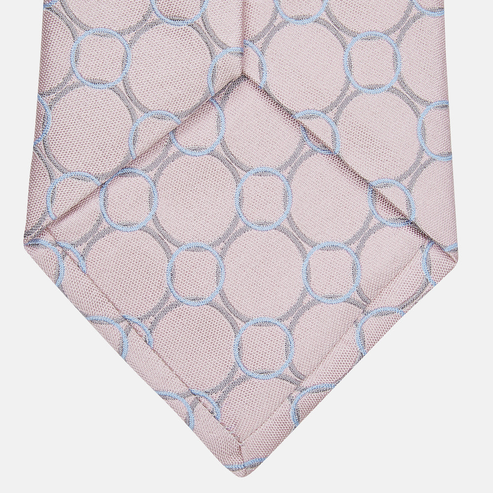 The Turnbull & Asser Baby Pink and Blue Circle Jacquard Silk Tie features a subtle geometric pattern, elegantly rolled against a plain white background.