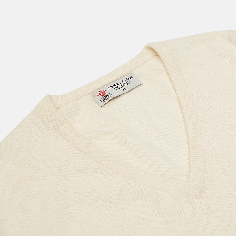 The Turnbull & Asser Cream Cashmere V-Neck Jumper is a luxury knitwear piece with ribbed cuffs and hem, shown laid flat on a white background. Its classic long-sleeve style offers timeless elegance.