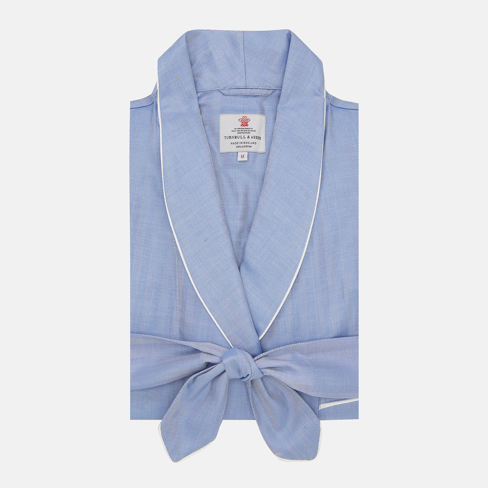 The Blue Herringbone Superfine Cotton Gown by Turnbull & Asser is a folded light blue robe with white piping and a tied front belt, featuring a branded collar label, displayed against a plain white background.
