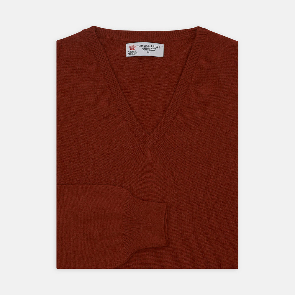 The Turnbull & Asser Harissa V-Neck Cashmere Jumper is shown folded in deep red with ribbed cuffs and a visible collar label, set against a white background.
