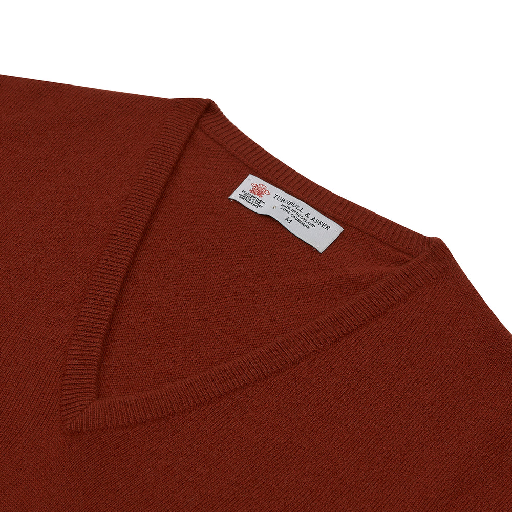 The Turnbull & Asser Harissa V-Neck Cashmere Jumper is shown folded in deep red with ribbed cuffs and a visible collar label, set against a white background.