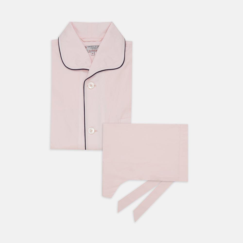The Pink Sea Island Cotton Pyjama Set by Turnbull & Asser features a folded light pink top with black piping and white buttons, shown beside a matching headscarf with two ties, highlighting luxury men’s sleepwear on a white background.