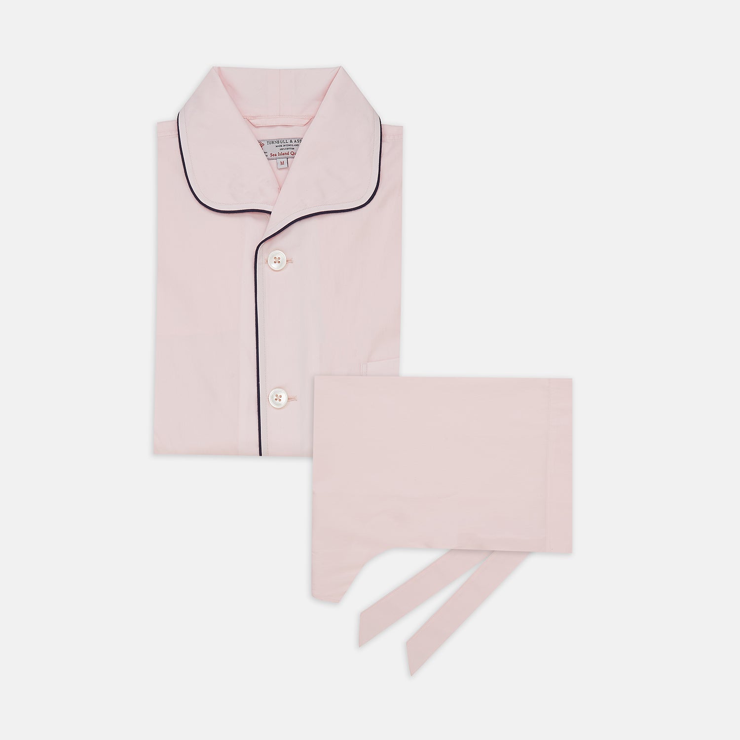 View 1 of product The Pink Sea Island Cotton Pyjama Set by Turnbull & Asser features a folded light pink top with black piping and white buttons, shown beside a matching headscarf with two ties, highlighting luxury men’s sleepwear on a white background.