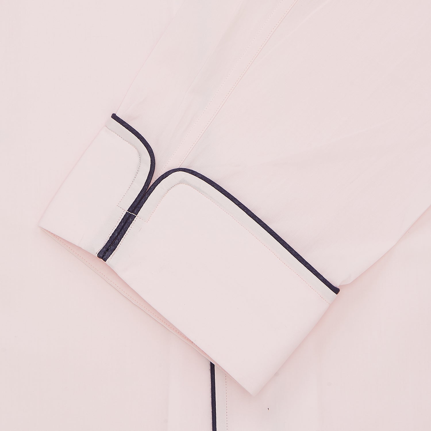 View 2 of product The Pink Sea Island Cotton Pyjama Set by Turnbull & Asser features a folded light pink top with black piping and white buttons, shown beside a matching headscarf with two ties, highlighting luxury men’s sleepwear on a white background.