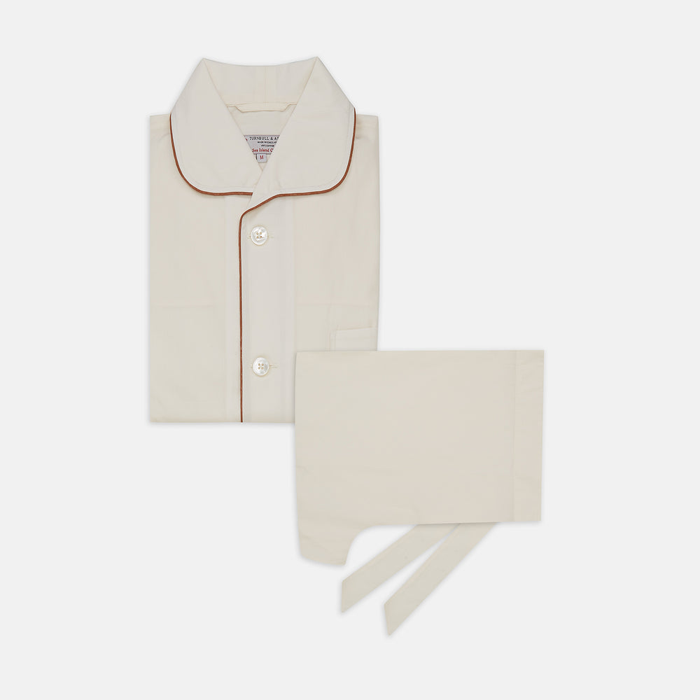 The Turnbull & Asser Cream Sea Island Cotton Pyjama Set features folded cream pyjamas with brown piping and a matching sleep mask with fabric ties, all elegantly displayed against a plain white background.