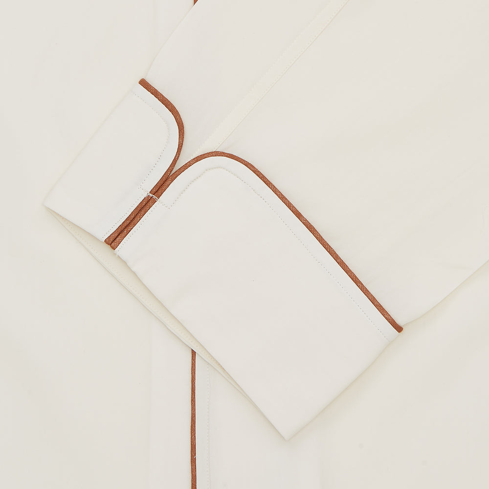 The Turnbull & Asser Cream Sea Island Cotton Pyjama Set features folded cream pyjamas with brown piping and a matching sleep mask with fabric ties, all elegantly displayed against a plain white background.