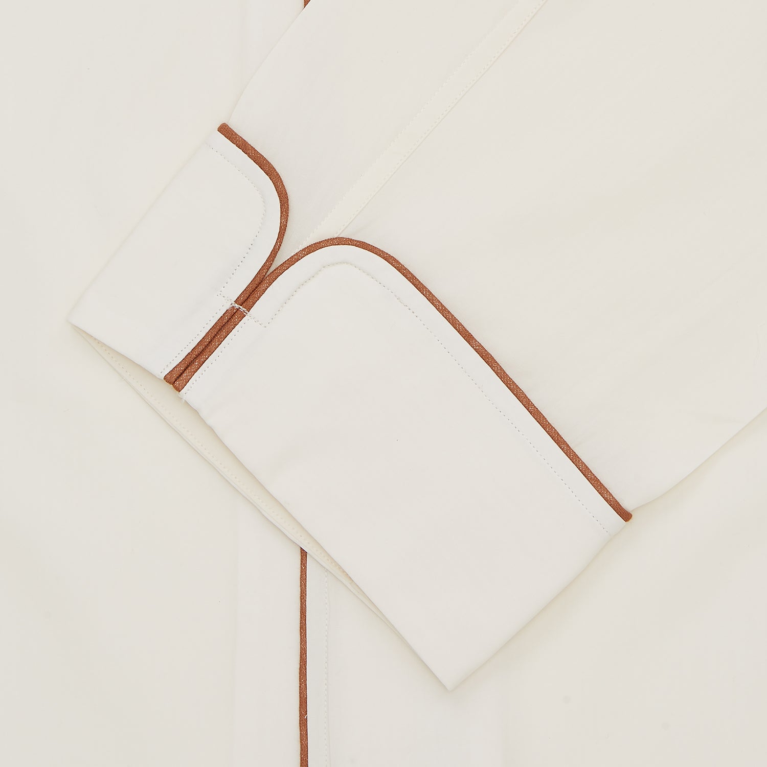 View 2 of product The Turnbull & Asser Cream Sea Island Cotton Pyjama Set features folded cream pyjamas with brown piping and a matching sleep mask with fabric ties, all elegantly displayed against a plain white background.