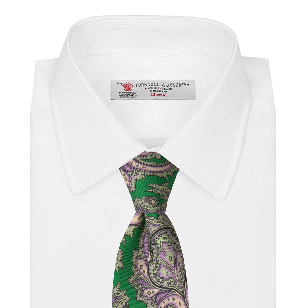 The Turnbull & Asser Green and Purple Large Paisley Printed Silk Tie features a light purple and white intricate paisley pattern, rolled up and set against a plain white background for timeless style.