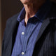 Close-up of a person wearing the Turnbull & Asser Blue Stripe Weekend Fit Nevis Shirt with Derby Collar and 1-Button Cuffs under a dark pinstripe blazer, focusing on the collar and upper chest. The face is partially out of frame.