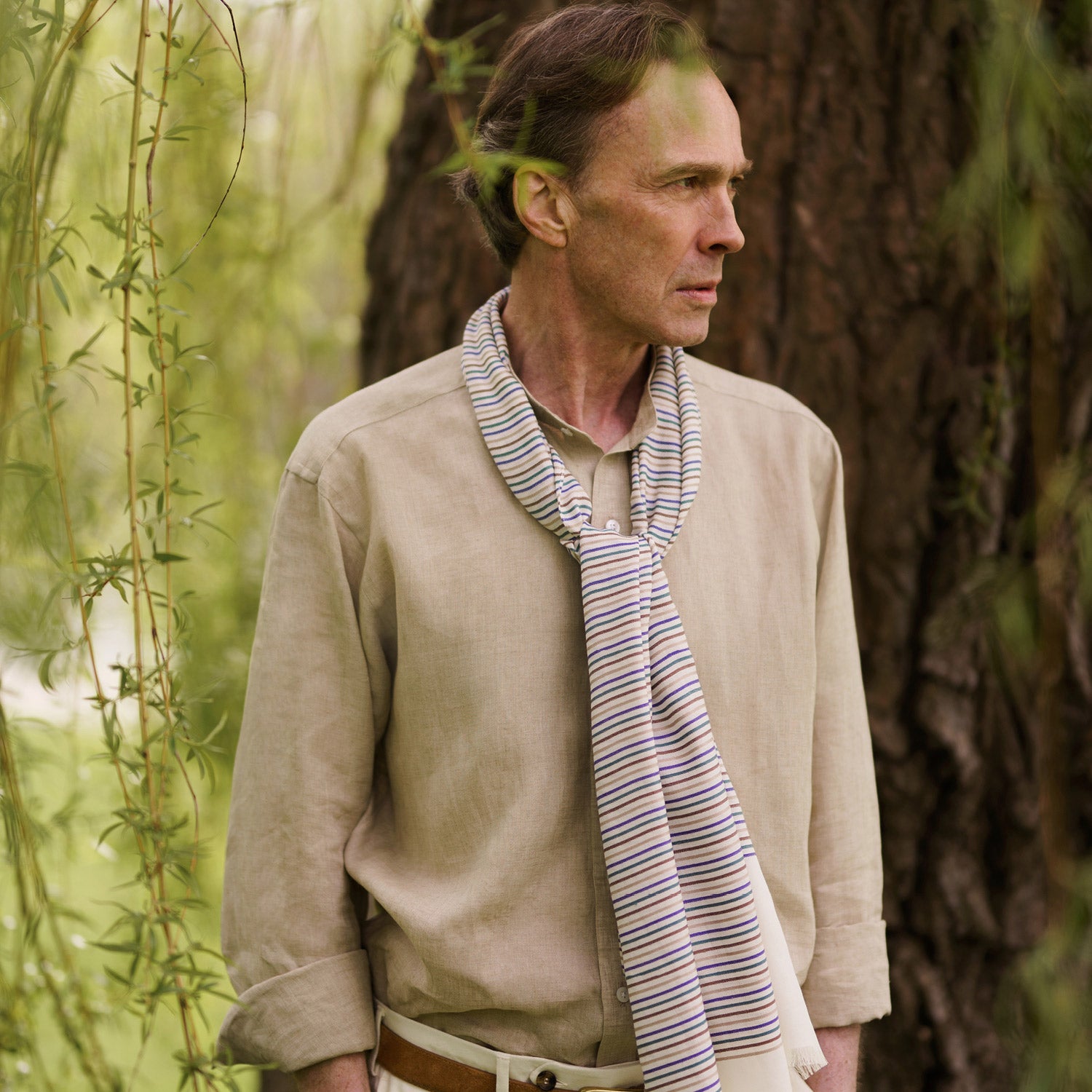 View 1 of product A man wearing a Turnbull & Asser Cream Abstract Stripe Print Lightweight Wool Scarf and a beige shirt stands outside by a tree, surrounded by greenery, gazing thoughtfully to the side.