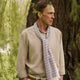 A man wearing a Turnbull & Asser Cream Abstract Stripe Print Lightweight Wool Scarf and a beige shirt stands outside by a tree, surrounded by greenery, gazing thoughtfully to the side.