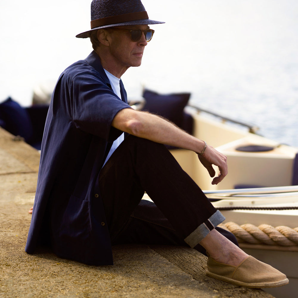 A man stands against a beige backdrop, wearing the Turnbull & Asser Navy Holiday Fit Silk Phillips Shirt with Revere Collar and dark pinstripe pants. He gazes downwards with a relaxed expression, embodying effortless summer style.