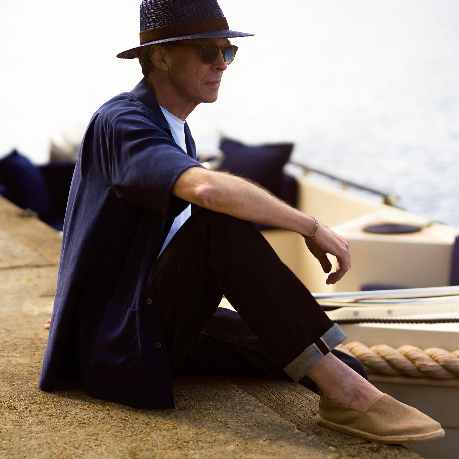 View 7 of product A man stands against a beige backdrop, wearing the Turnbull & Asser Navy Holiday Fit Silk Phillips Shirt with Revere Collar and dark pinstripe pants. He gazes downwards with a relaxed expression, embodying effortless summer style.