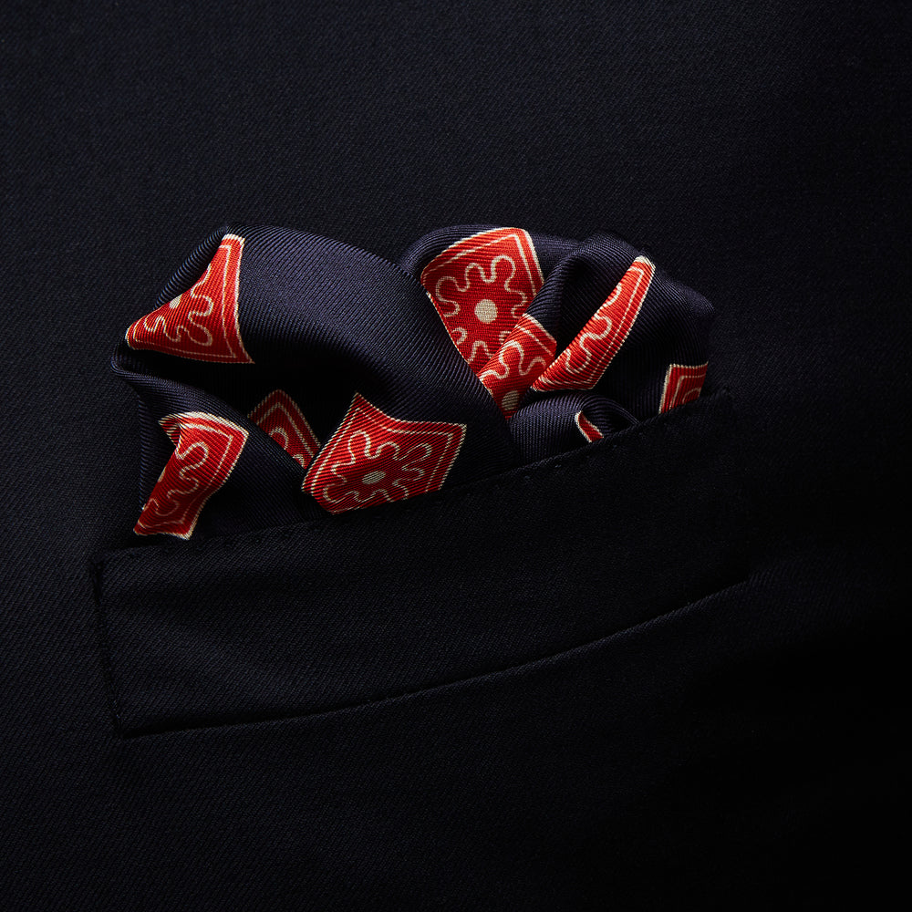The Navy and Cherry Red Tiled Floral Silk Pocket Square by Turnbull & Asser features a blue background with red and white geometric floral patterns, a decorative border, and the brand's name in one corner.