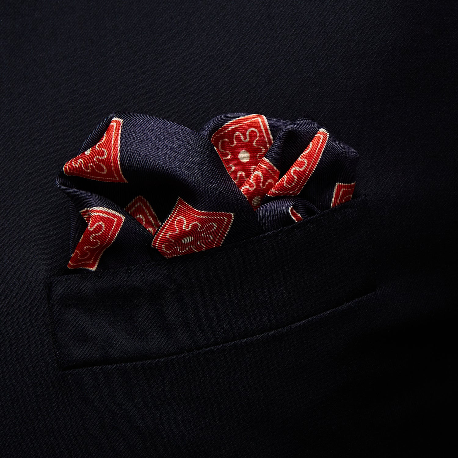 View 3 of product The Navy and Cherry Red Tiled Floral Silk Pocket Square by Turnbull & Asser features a blue background with red and white geometric floral patterns, a decorative border, and the brand's name in one corner.