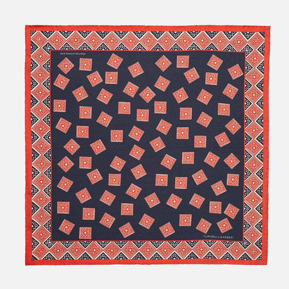 The Navy and Cherry Red Tiled Floral Silk Pocket Square by Turnbull & Asser features a blue background with red and white geometric floral patterns, a decorative border, and the brand's name in one corner.